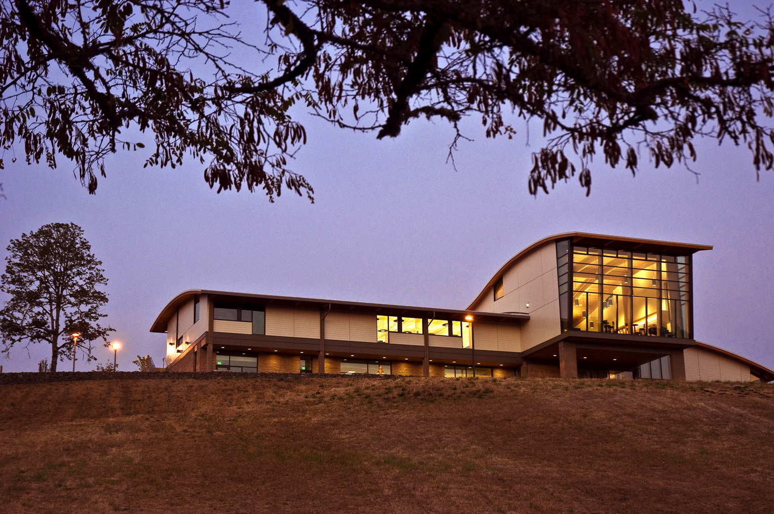 Umpqua Community College Danny Lang Center. Perched high on a hill and captured at dusk, this image shows a long, narrow winery and teaching building with a rounded roof. Portruding from the body of the building is a double-story tasting room which is lit from within.
