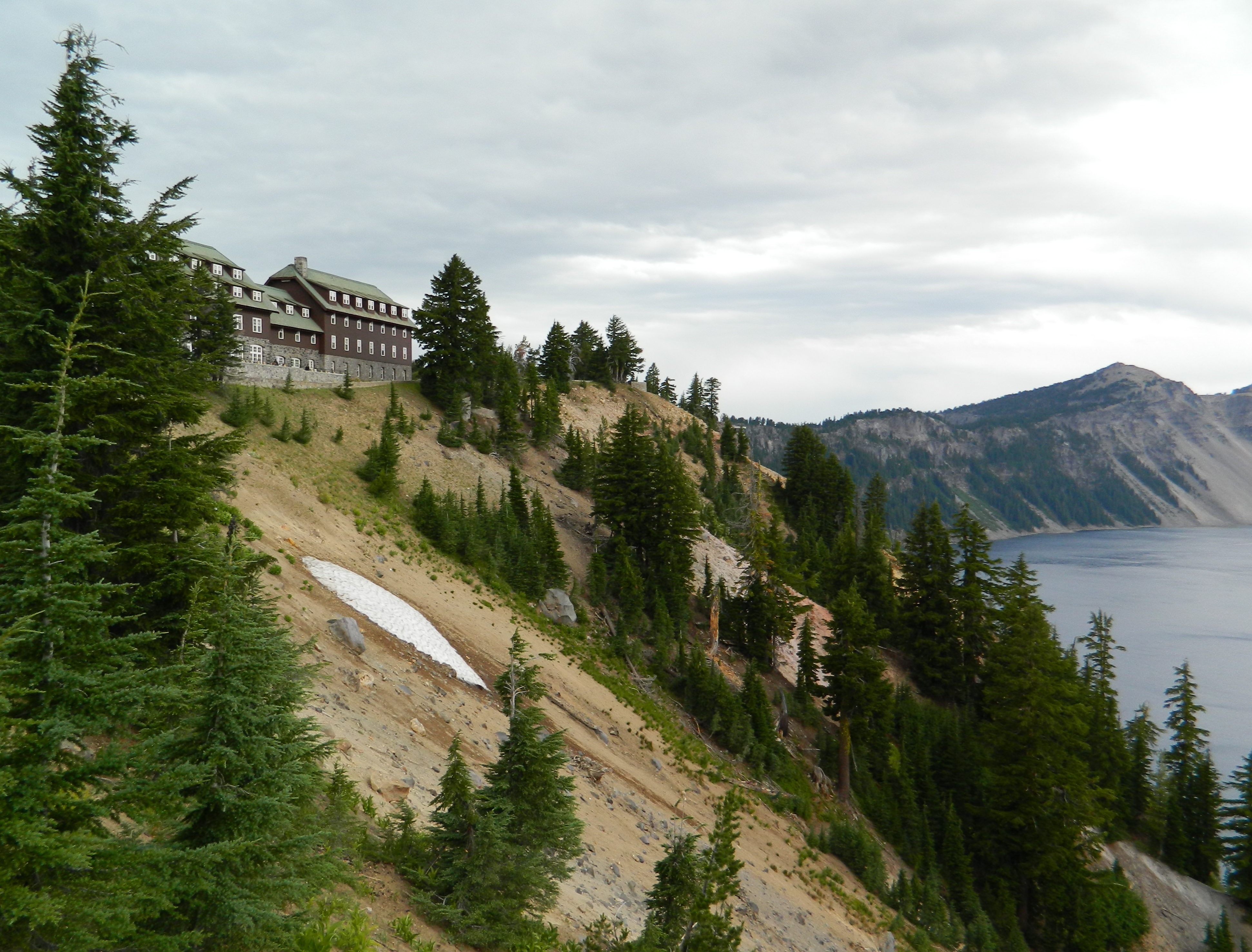 This image shows a sweeping view which includes the southernmost edge of crater lake with a steep, tree-filled slope leading from the lake up to a long, rustic four-story lodge. The first floor is clad in stone and the upper floors are wood-clad and painted deep brown. The pitched, shingled roof is a pale green.