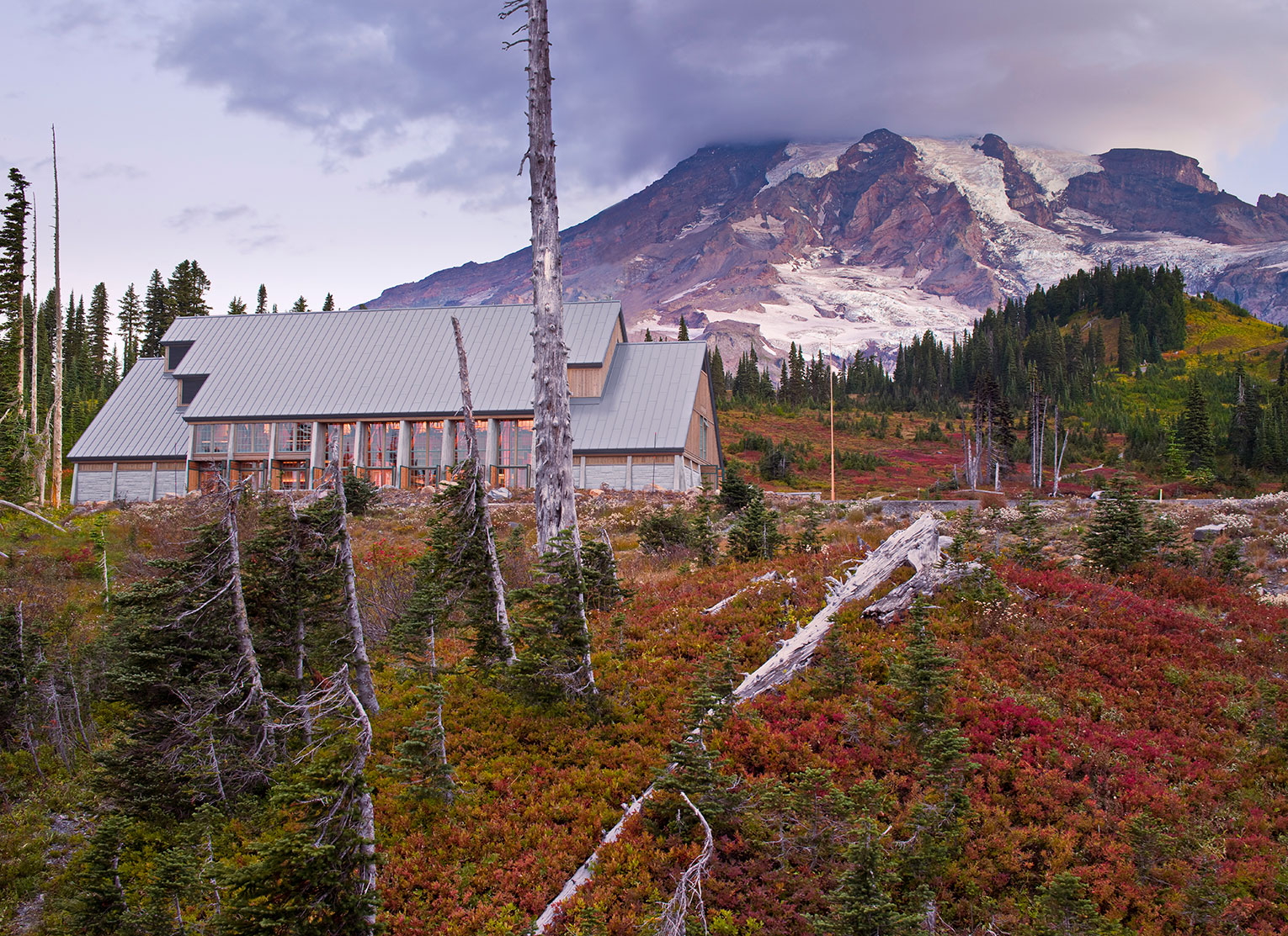 This image shows a large new visitor center with a steeply-sloped metal roof and floor-to-ceiling windows and doors which are lit from within. In the background, Mount Rainier looms large and its peak is partially covered by a large cloud. The sloping site is covered with evergreens, snags, and blanketed in red and yellow plants.