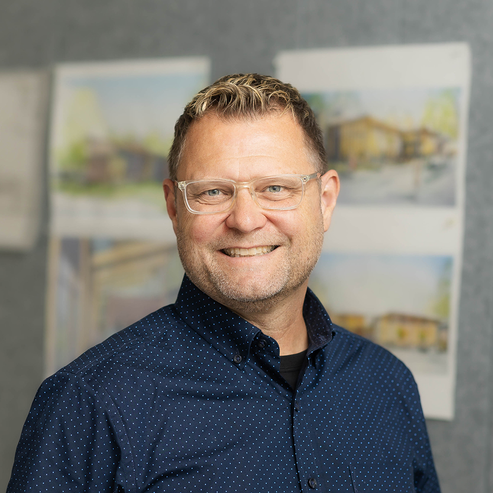 Professional headshot of a smiling man wearing glasses and a navy patterned shirt, standing in front of pinned architectural renderings