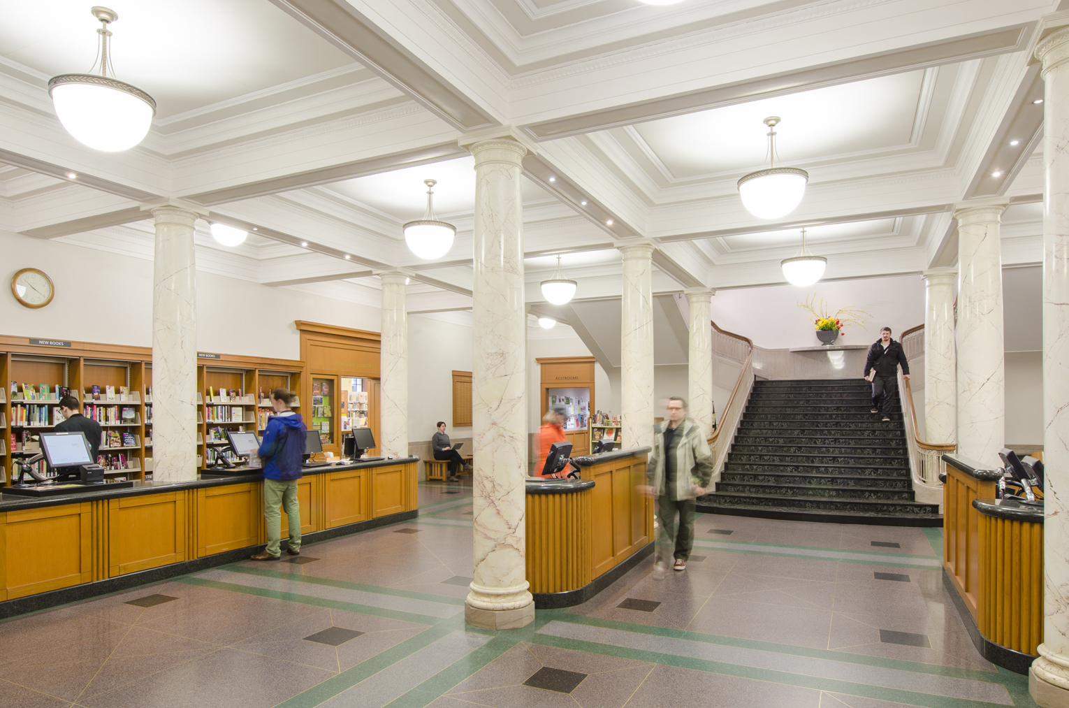 Multnomah County Central Library Rehabilitation. Large, open Georgian style library loby with marble columns flanking self check-out stationin the foreground and a dark marble grand staircase in the background. Box beam ceilings can be seen overhead and the floor is a simply patterned geometric terrazzo.