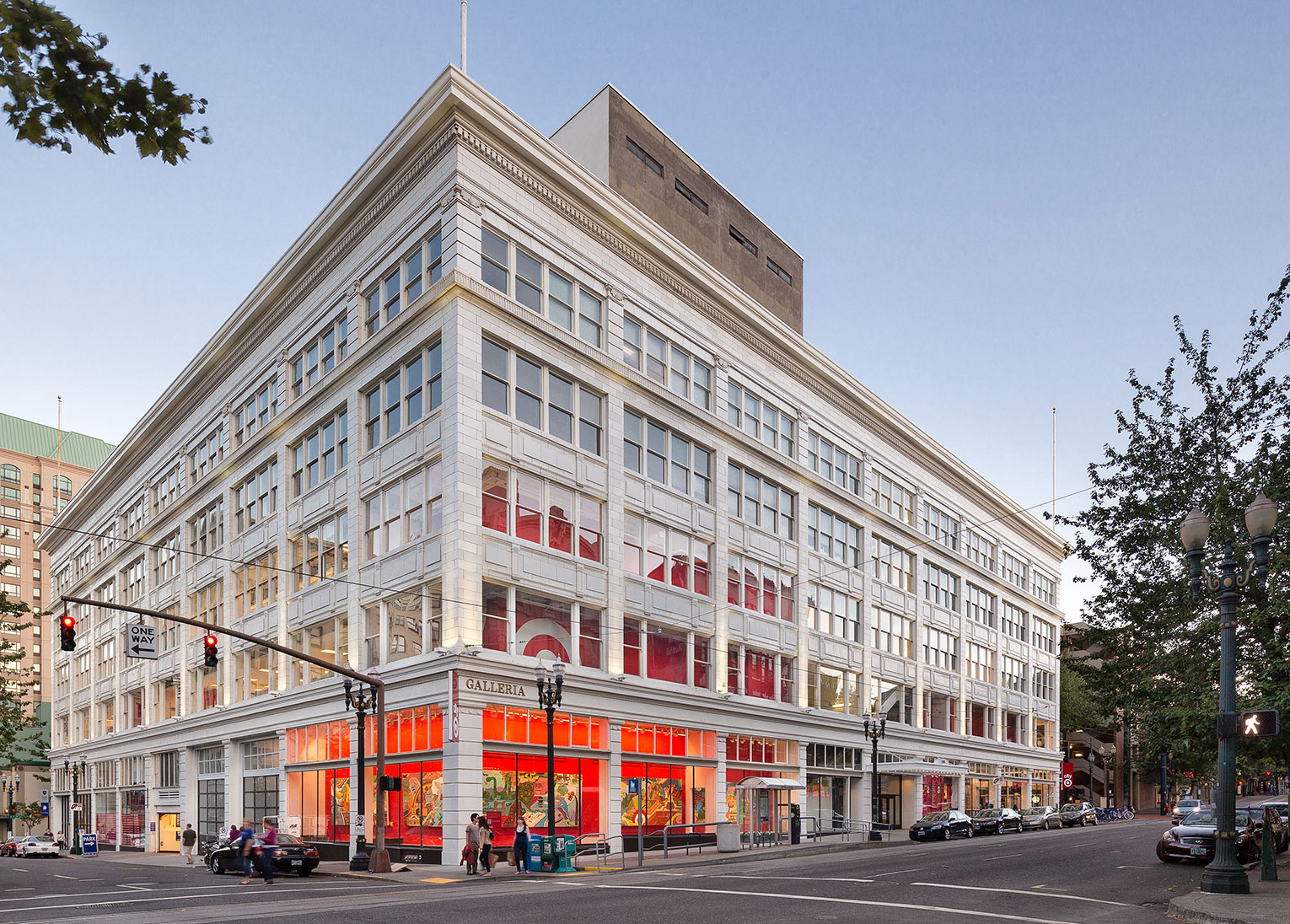 Corner view of downtown Portland's white terra cotta-clad historic department store, the Galleria Building, with many windows. The ground floor corner in the foreground is an art gallery with large, colorful paintings hanging on a bright orange wall. To the left of the gallery are two garage-style doors which appear to be loading docks. Through the windows of the second and third floors, a large Target logo can be seen, indicating the presence of the retailer in the space.