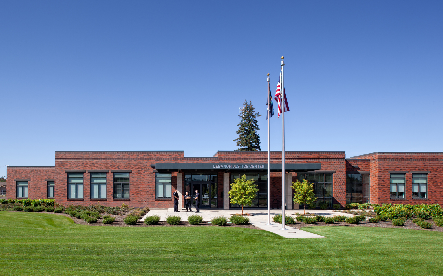 Lebanon Justice Center. Single-story, long, brick-clad public safety building with a small plaza in front, surrounded by a vast green lawn. Both the American and Oregon flags are being flown on a sunny, cloudless day.
