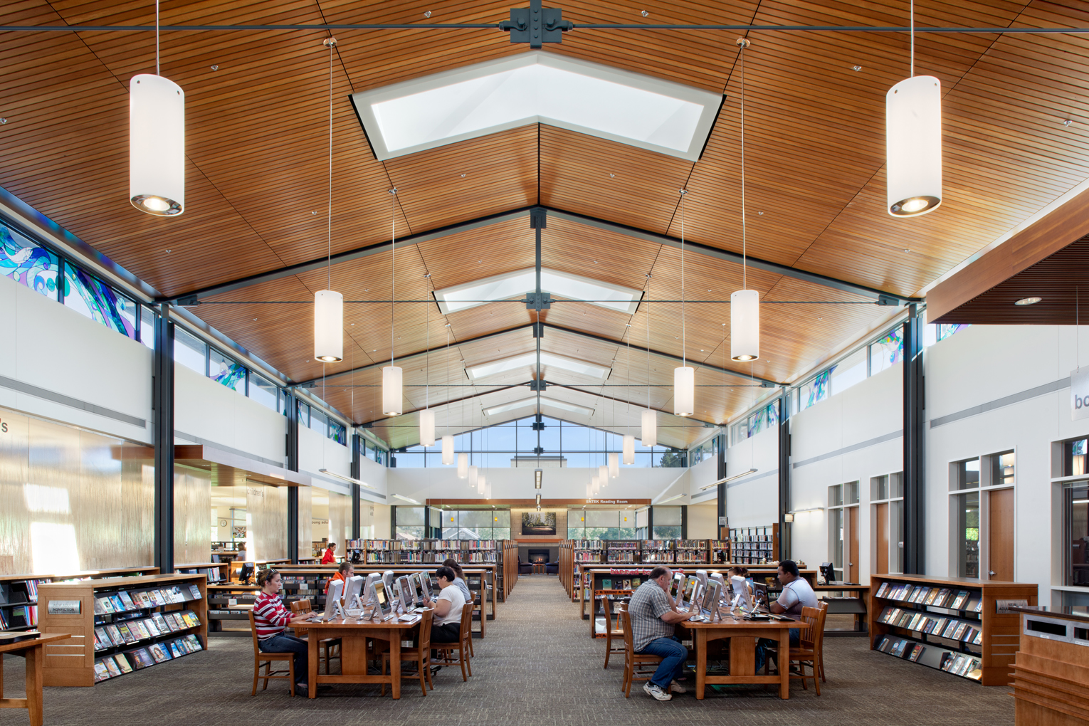Lebanon Public Library. A large, open library reading room with tables and computers in teh foreground and library collections in the background. The wood-clad ceiling is very high and features a pitched roofline and multiple skylights. There are stained glass clearstories along the sides of the roofline, and a triangular one made of transparent glass at the end. In the very back of te building a lit fireplace can be seen at the end of the aisle between stacks.