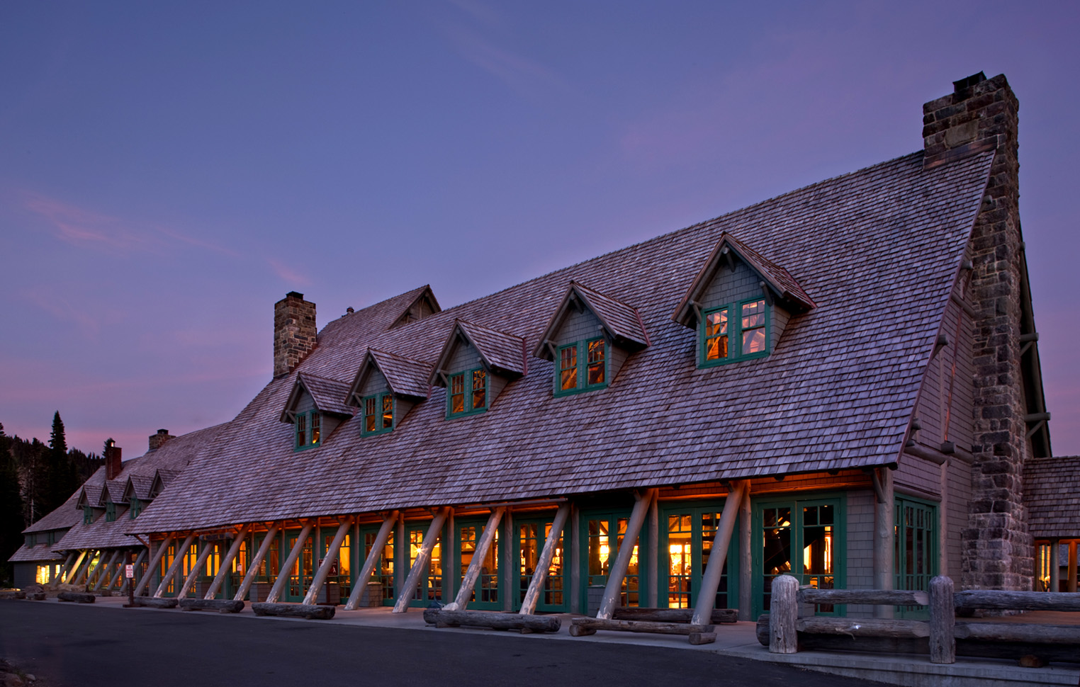 NPS Paradise Inn Rehabilitation. A large, rustic wood and log-framed lodge is shown at dusk while lights glow from within . Dormer windows line the roof, and a massive stone chimney is featured on the end of the building.