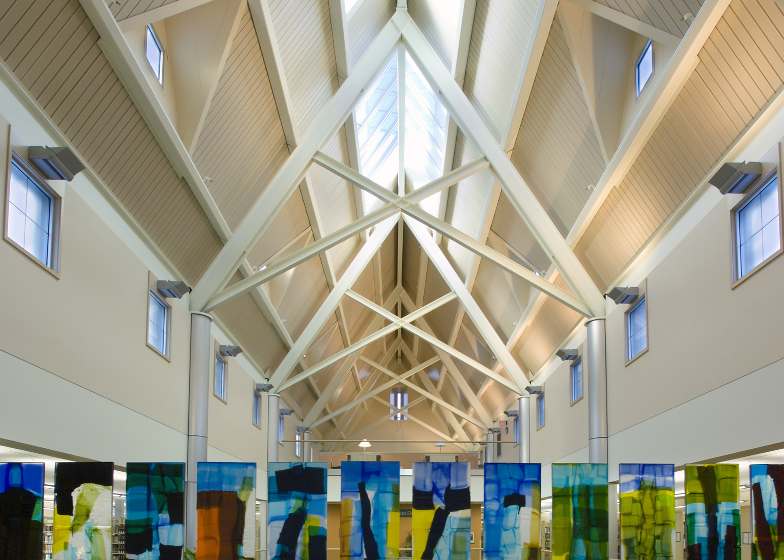 Pacific University Library. The foreground of this image shows the upper portion of a wall comprised of colorful fused glass panels. Both above and beyond the panels, a high, gabled ceiling with elegant white exposed support trusses enhances the grand cathedral-like space.