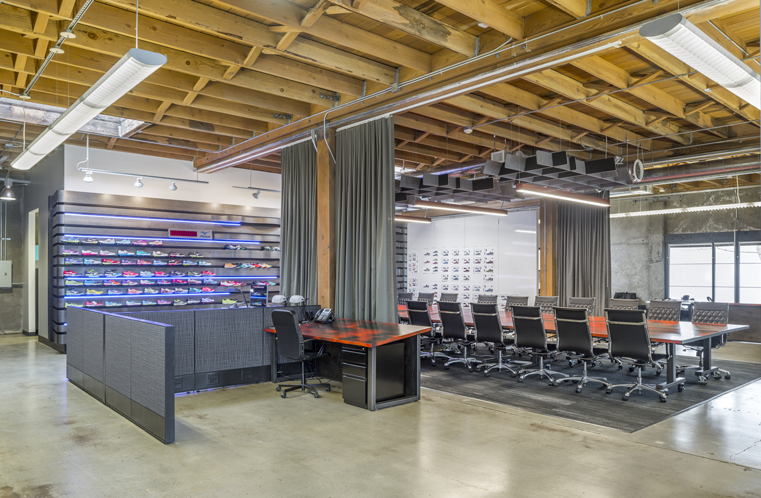Mizuno Sozo Design Studio. A large, open office with concrete floors and an exposed wood truss ceiling features a reception desk with brightly-colored prototypes of running shoes on display in the background. A large conference table with chairs is flanked by floor-to-ceiling curtains which can be pulled closed to surround the table to create a private conference room.