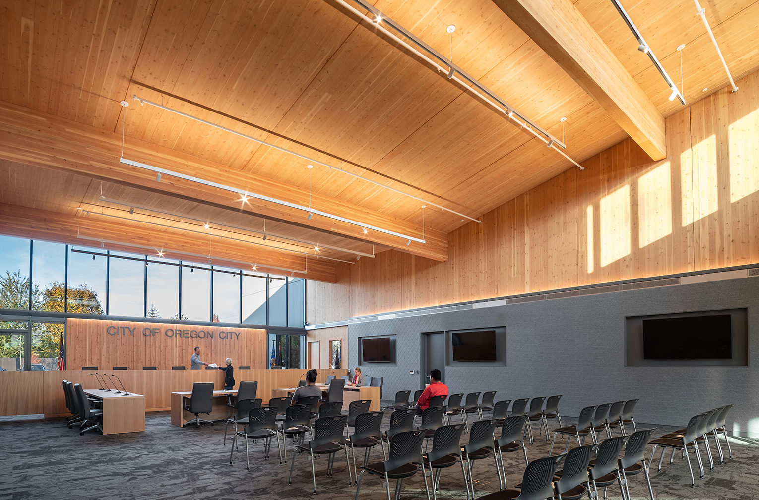 Robert Libke Public Safety and Municipal Courts Building. A large council chamber with a dias int he background and seating in in the foreground. The high ceiling is made of mass timber and sunlight streams through windows behind the dias, bathing the room in natural light.