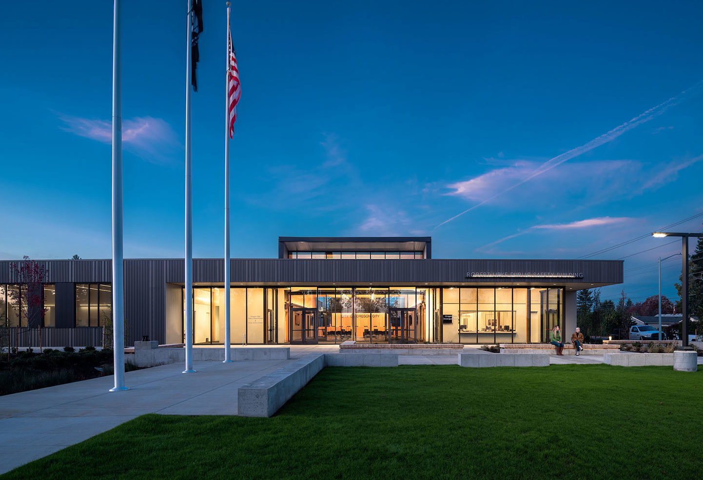 Evening image of lawn and open plaza in front of the single-story Robert Libke Public Safety and Municipal Courts Building which reveals the mass timber interior of the lobby and council chambers,