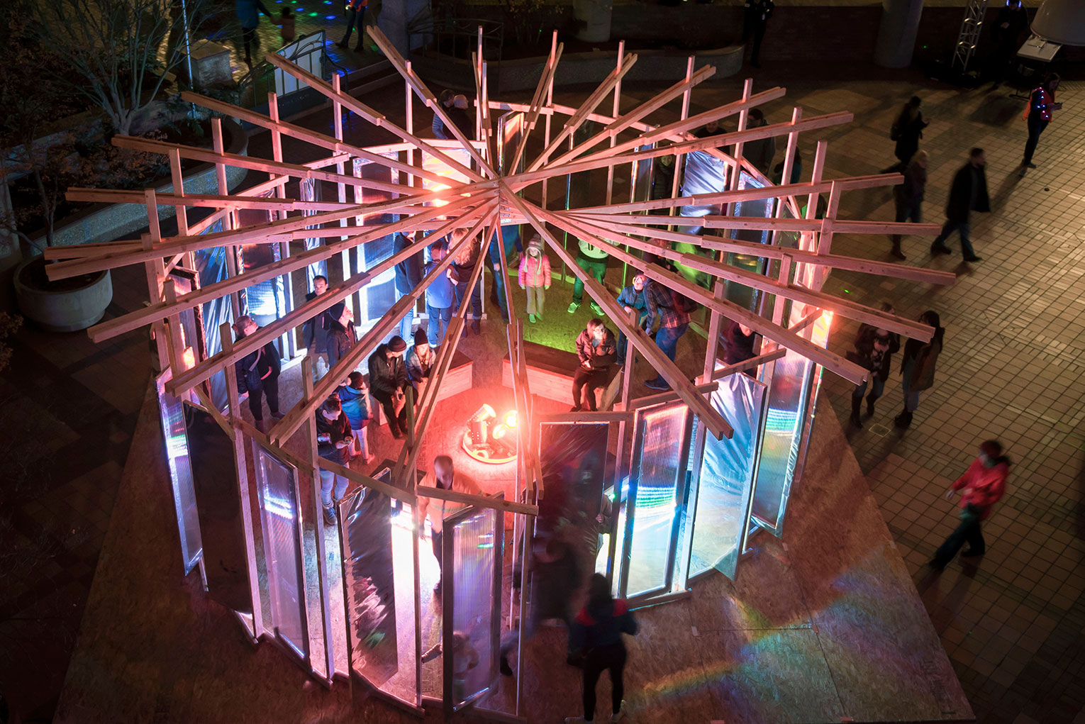 Winter Lights Festival: People to People Through Light. Aerial vew of multi-colored interactive light display topped with fanned out wooden beams. Image shows people moving in and through the structure.