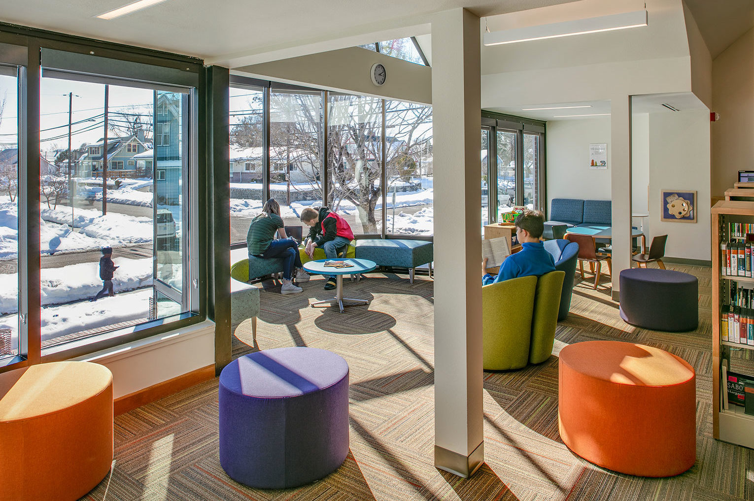 Goldendale Community Library Interior Renovations. Brightly daylit children's area in a library filled with colorful furniture and a rounded window seat. Snow on the grounds and street surrounding the building.