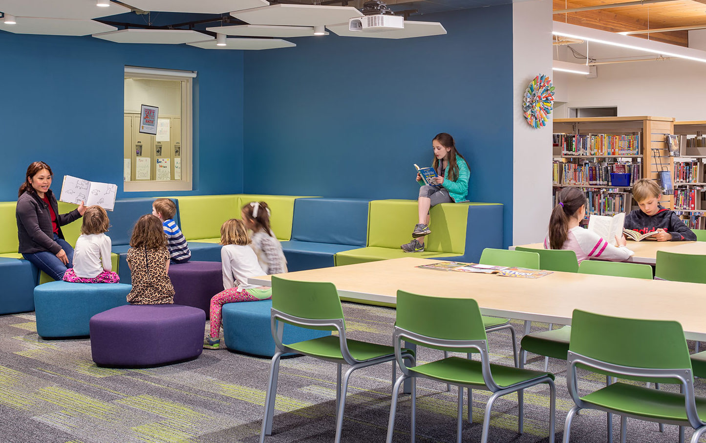 Portland Jewish Academy Learning Commons. Children sit in brightly-colored storytime area while a teach reads aloud to them. Blue walls surround the space and hexagon acoustic baffles hang from ceiling. Long reaing tables are featured in the foreground and book shelves in the background.