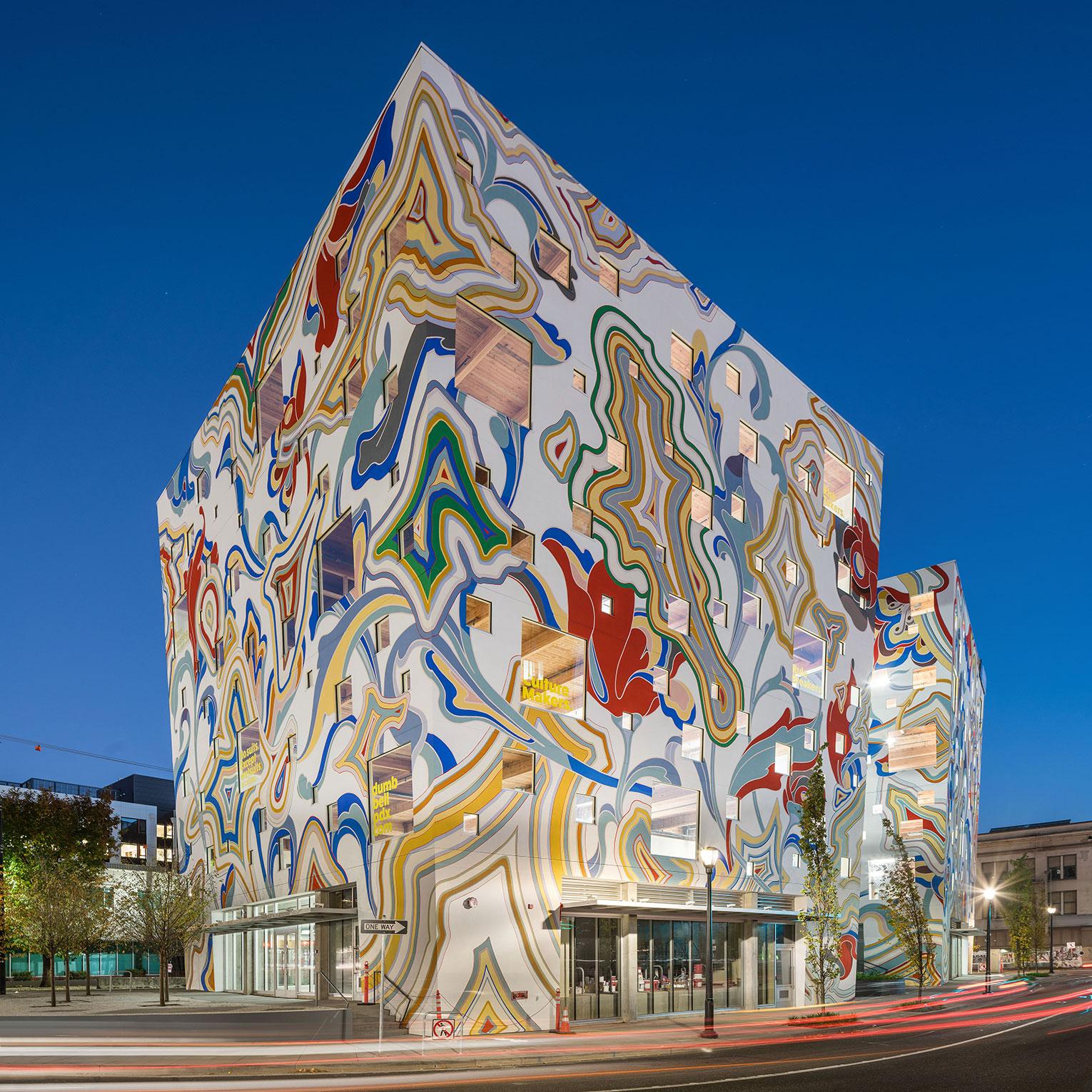 Dusk shot of the Fair-Haired Dumbbell. The six-story building is seen from the corner and the windows are of various sizes and are randomly placed. The exterior is completely covered with a colorful swirling mural abstractly representing flowers, geodes and leaves.