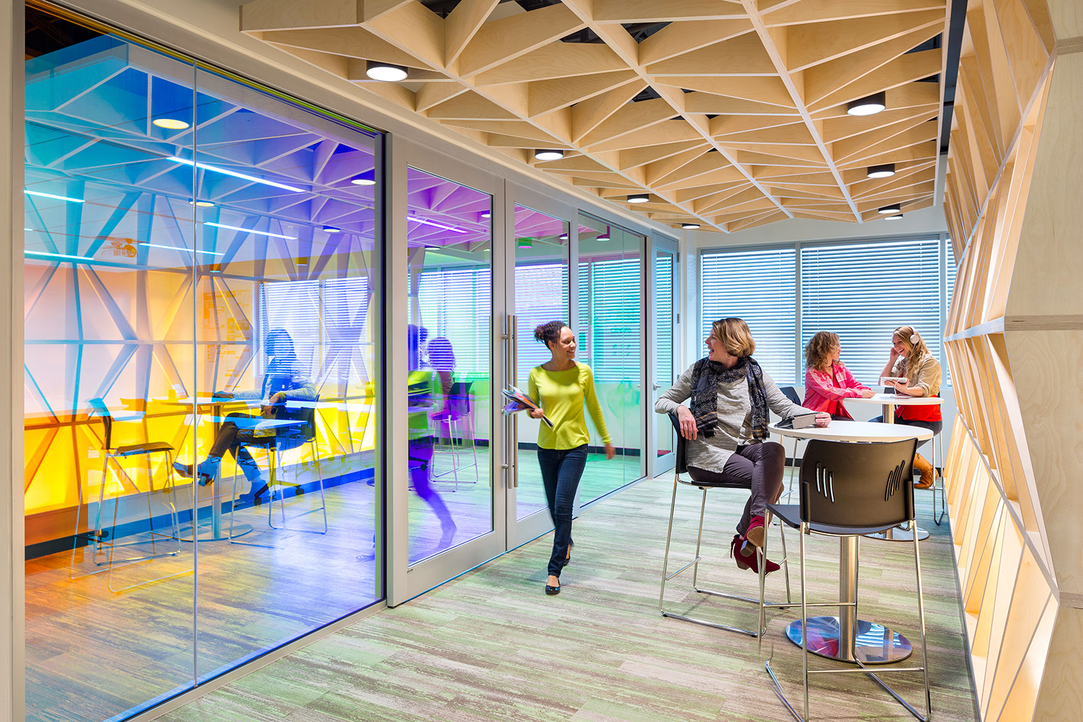 CD Baby Headquarters. Hallway with bistro-type tables for collaboration or quiet work. Multi-colored light reflects off sliding glass doors on the left. Ceiling and right-side wall are covered with custom woodwork in a geometric pattern. Two women can be seen visiting at the table in the background, and one woman sits at the table in the foreground, greeting a passing co-worker.