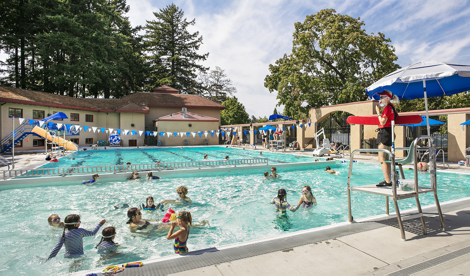 Peninsula Park Pool. A sunny summer's day at a bright blue outdoor pool featuring several children playing in the water while a lifeguard supervises from a tall chair on the pool deck.