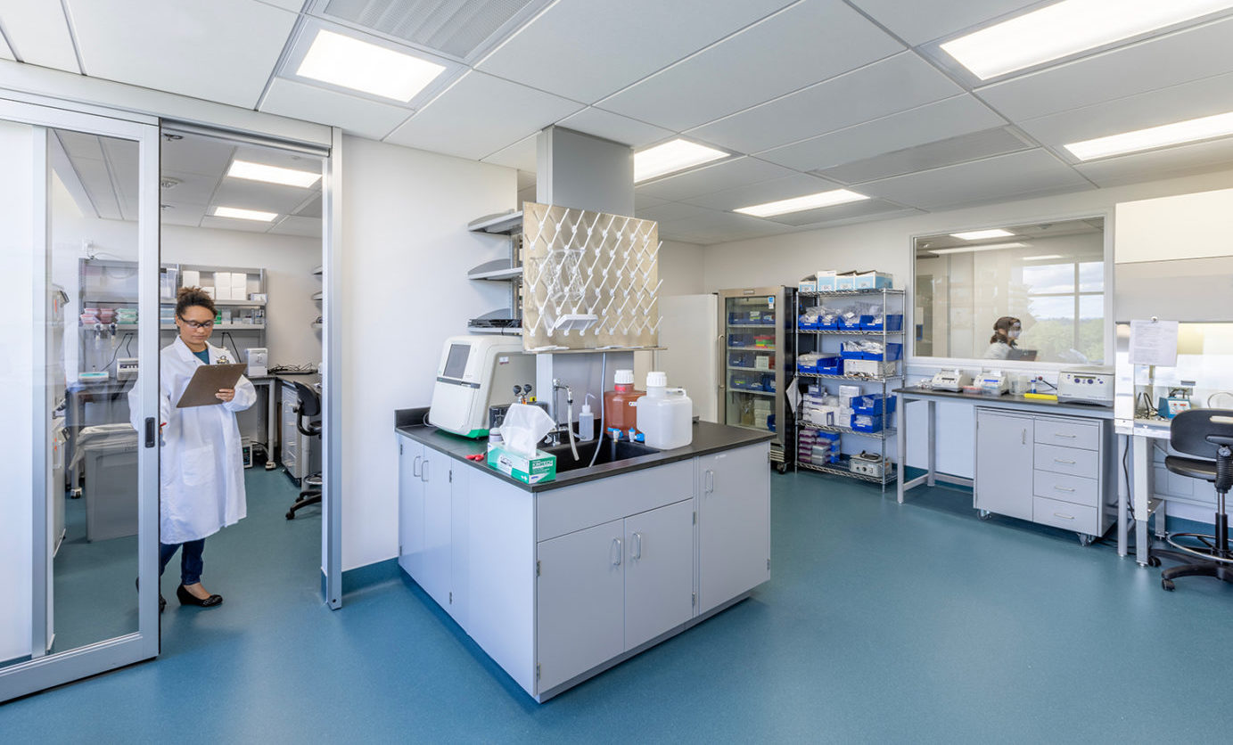 Vir Biotechnology Office and Laboratory. An African American woman in a lab coat, carrying a clipboard enters a brightly-lit, renovated lab with grey cabinets and turquoise flooring..