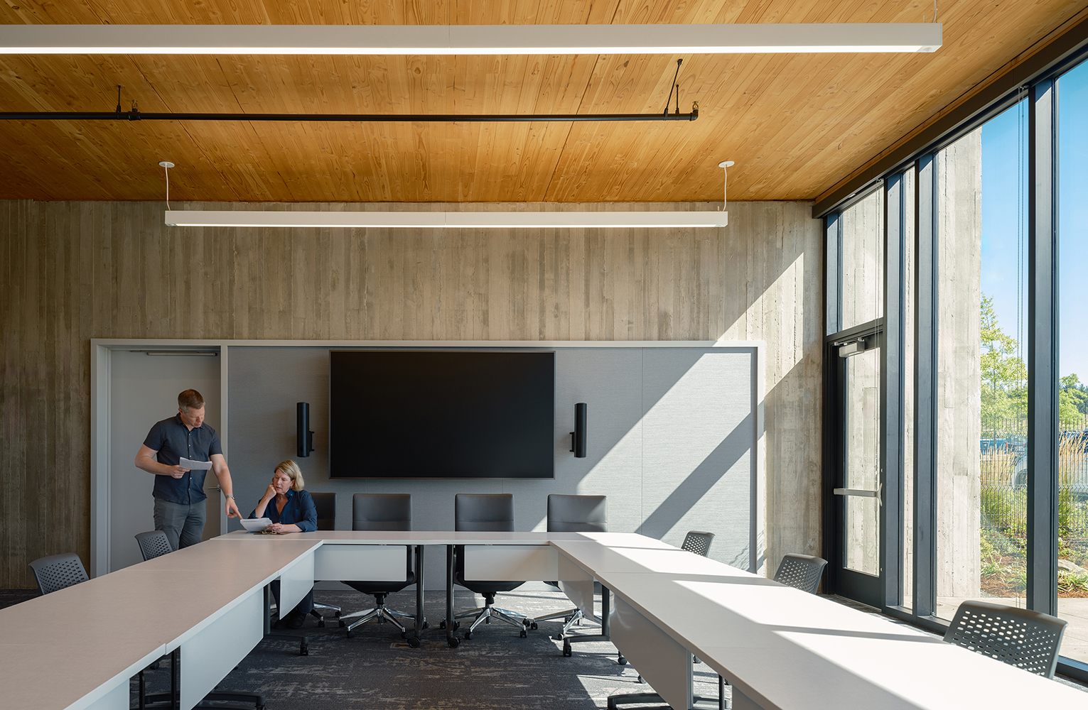 Two staff review documents in a daylight-filled boardroom featuring a CLT ceiling, boardroom technology wall, and floor-to-ceiling glazing overlooking the landscape.