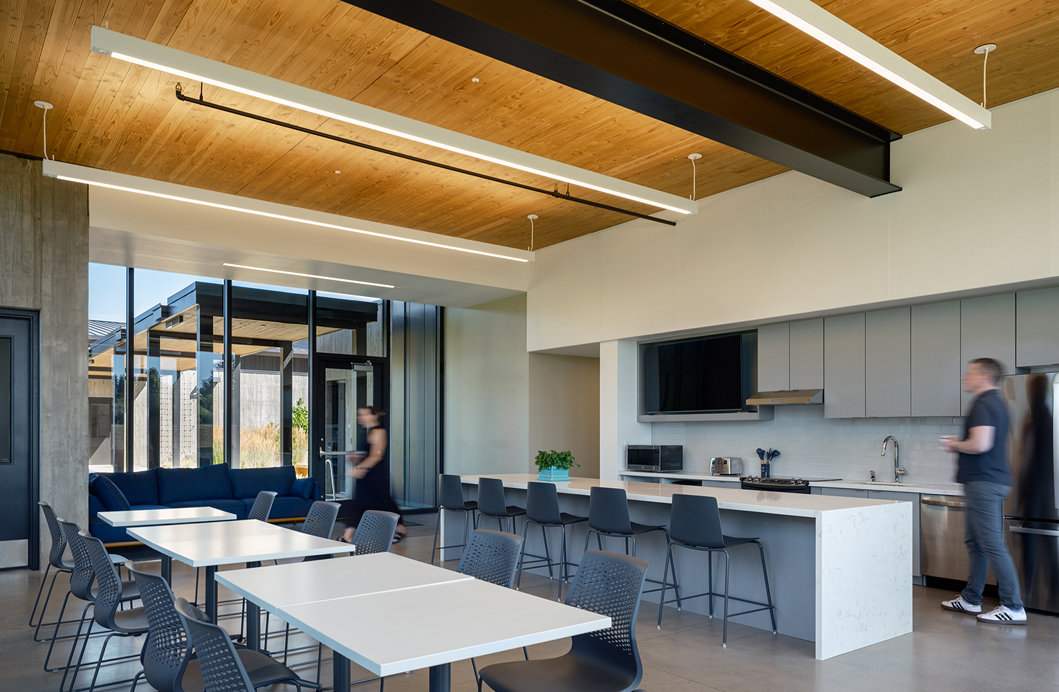 Staff walk through a bright break room featuring a CLT ceiling, large windows to the courtyard, and an open kitchen with communal seating.