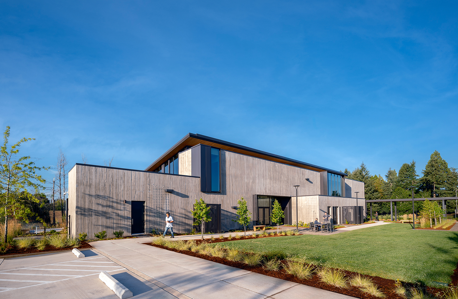 Staff walk through the Sunrise Water Authority courtyard beside the administrative building, where board-formed concrete walls, tall windows, and new plantings frame a sunny outdoor gathering lawn.
