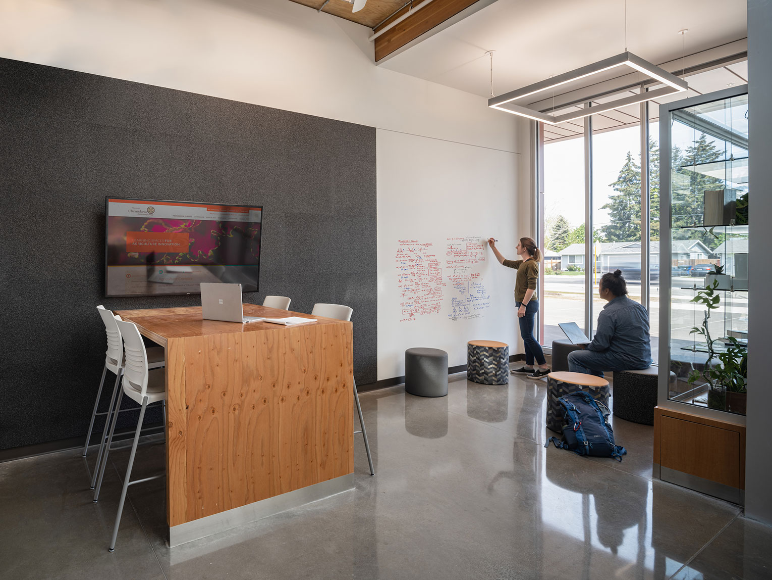 Woman making writing on an erasable wall, while man sits and takes notes on laptop