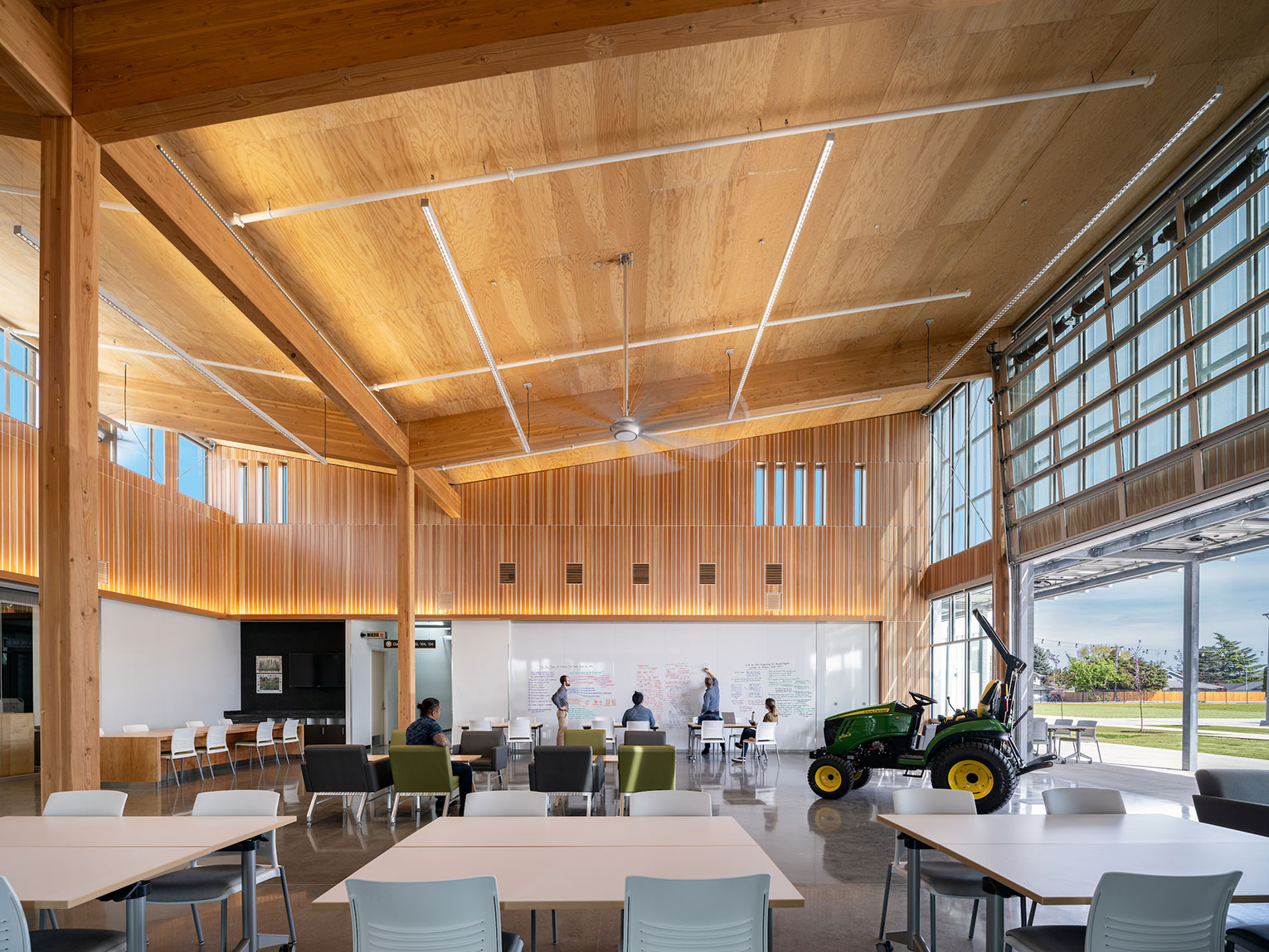 Large communal space with CLT ceiling and walls and roll-up windows. People writing on an erasable wall in background