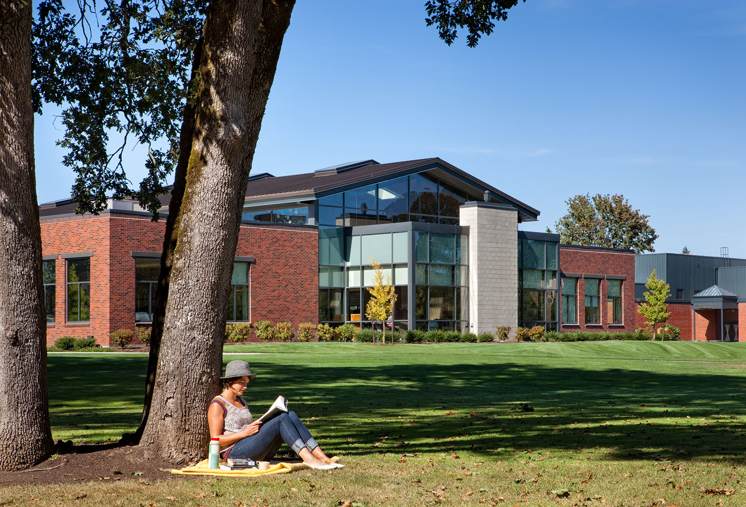 Woman sitting under tree reading in foreground, with single-story library in background