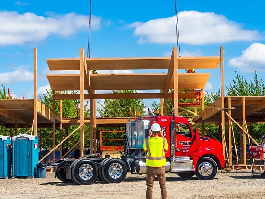 A,am om a yellow safety vest as seen from behind, facing the red cab of a semi truck and the shell of a building under construction