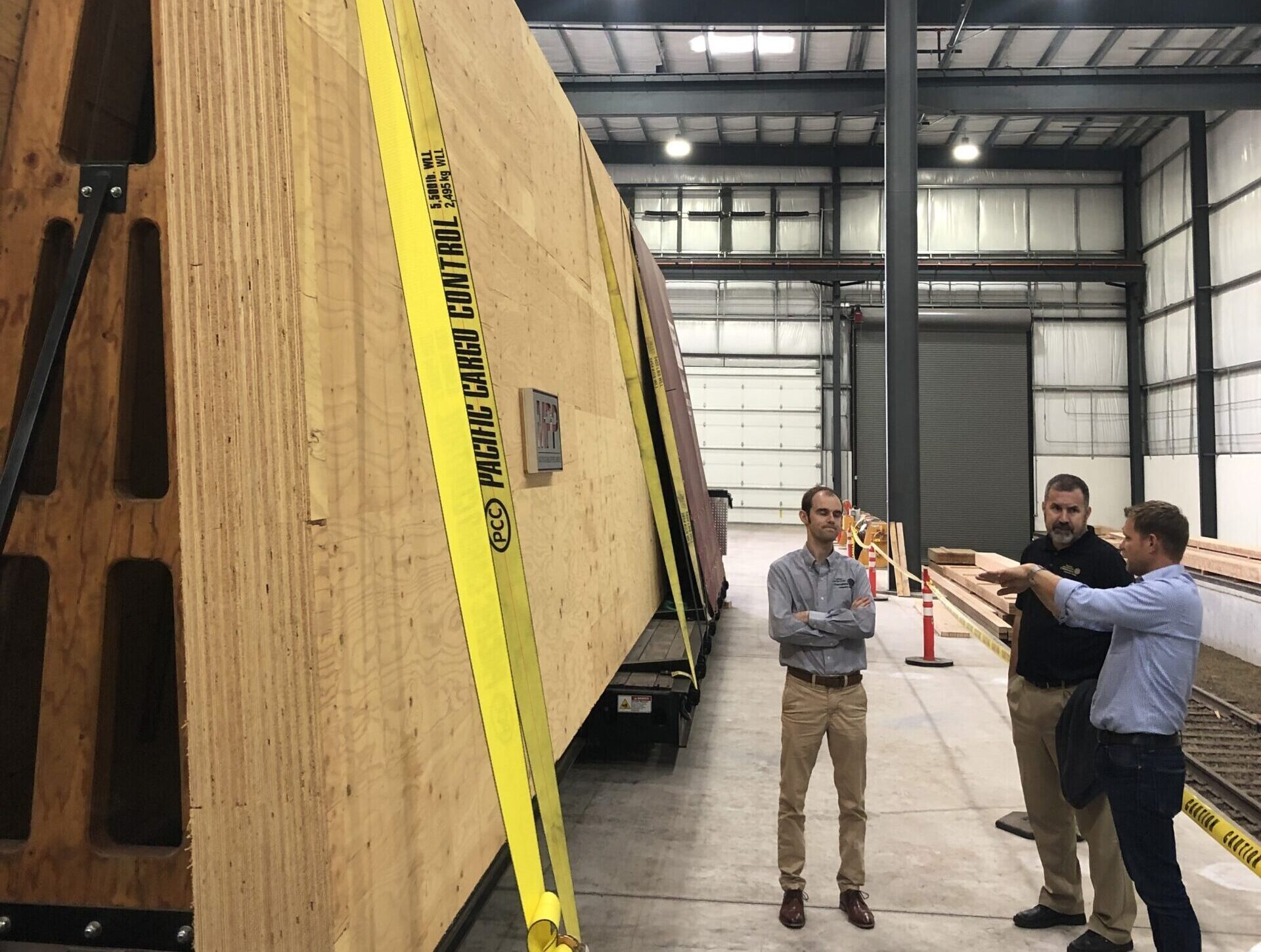 Three men in a warehouse looking at a massive mas timber panel that is strapped down.