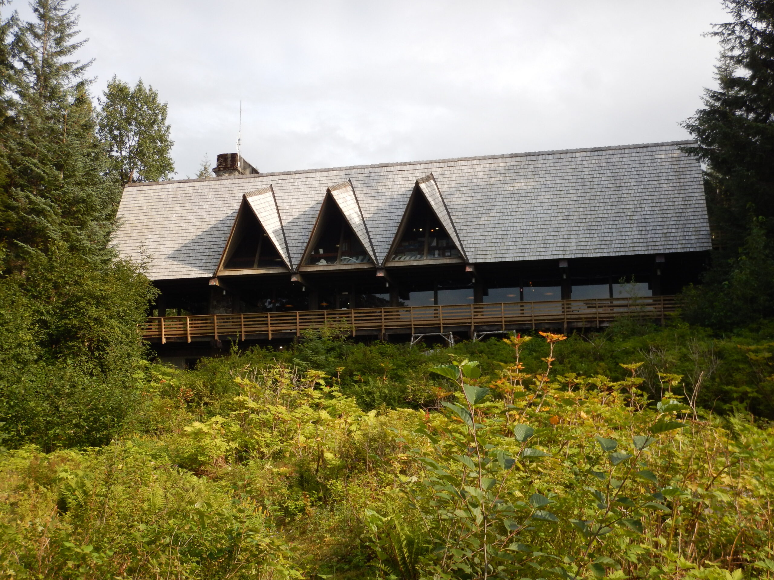 A long, lodge building surrounded by trees and vegetation. Three peaked dormer windows sprout from the roof