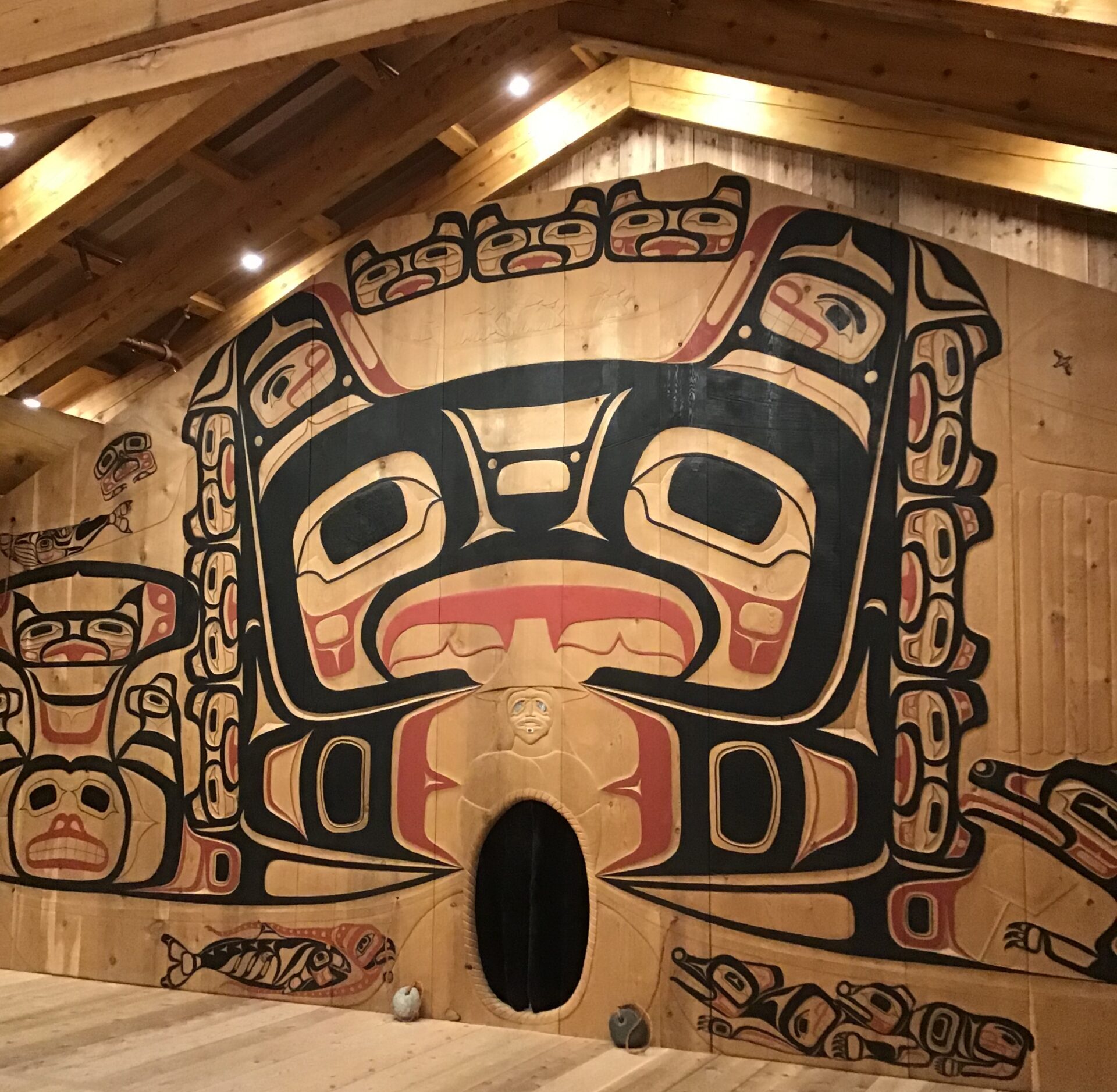 A large room with wood floors, and a beamed wooden ceiling. The mural painted on the wood wall depicts Native American symbols