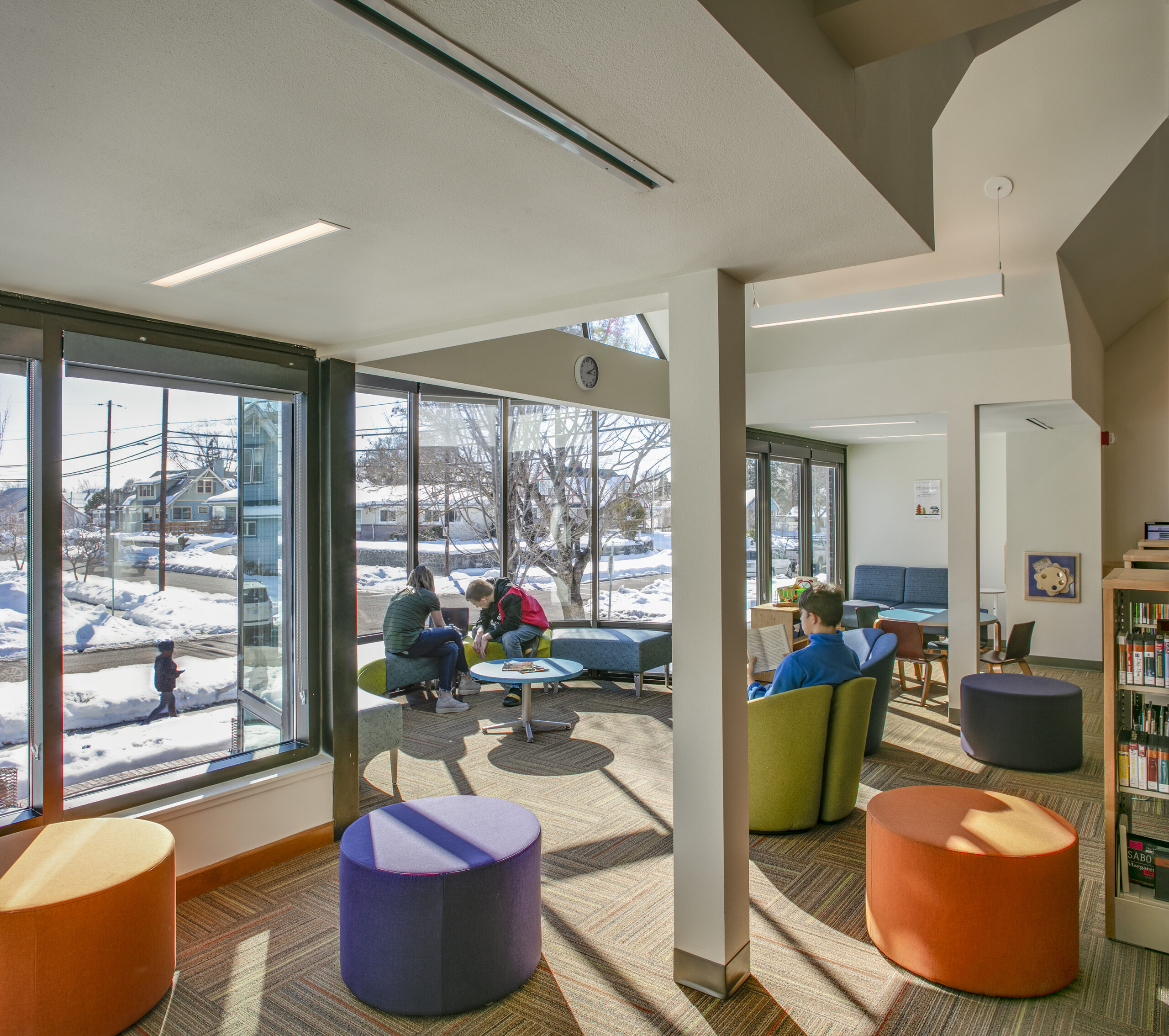 A brightly furnished children's area in a library. Several children are sitting down reading in front of a wall of windows which look out to the snowy surrounding neighborhood
