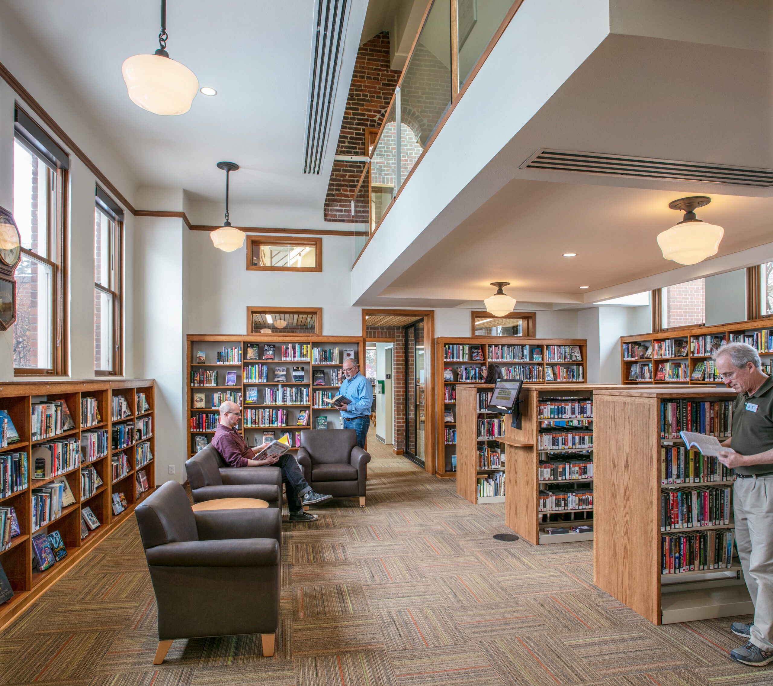 A small library reading room one man sits reading on an upholstered chair, and two others are standing, flipping through books