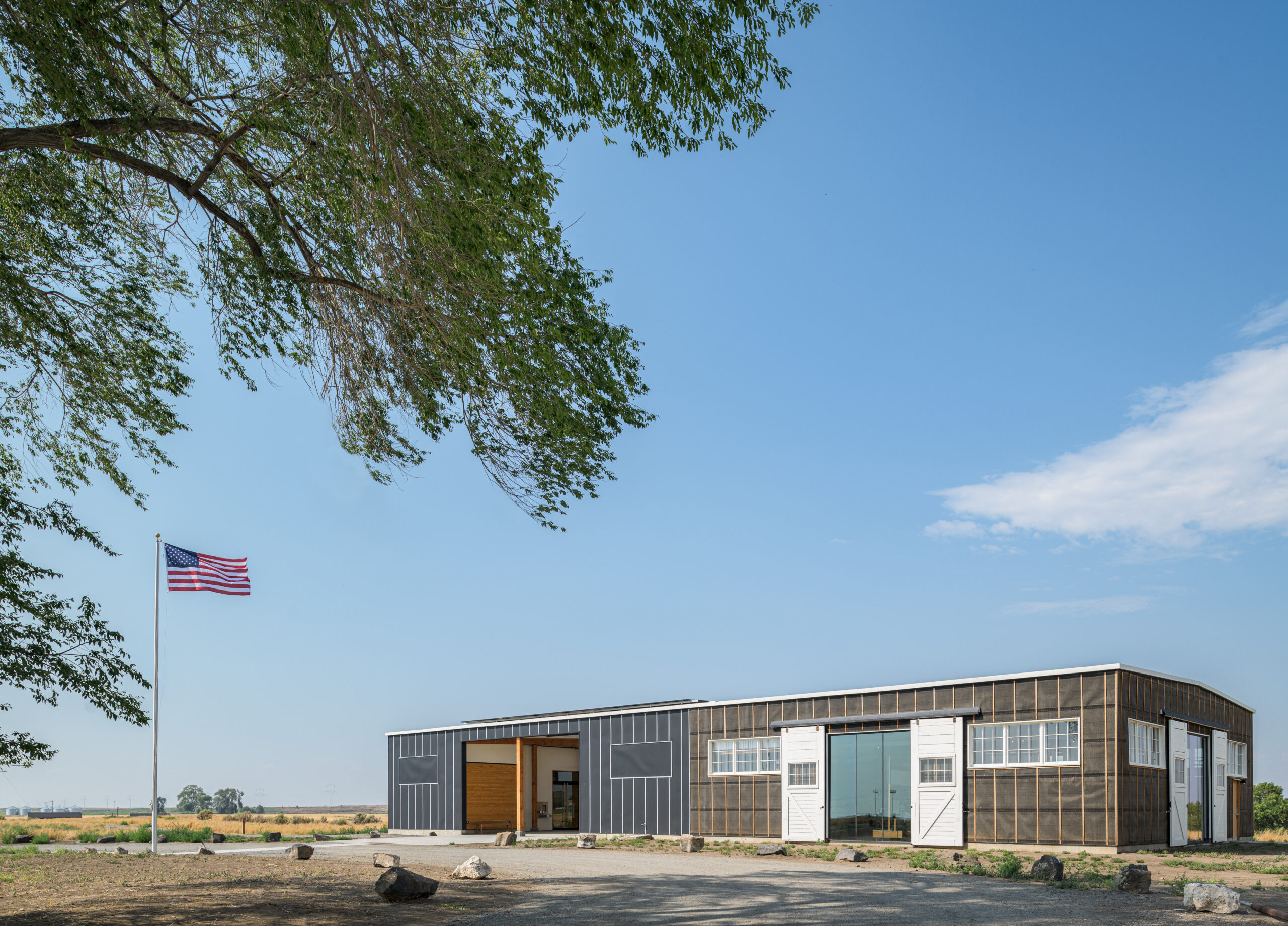 A long. narrow visitor contact station on a barren Idaho site. Photo is framed by a tree, and an American flag is flying in the background against a blue sky