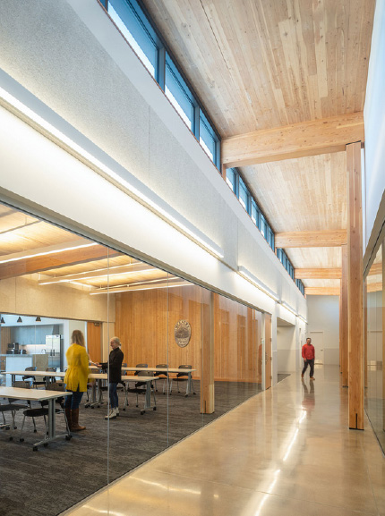 A man in a red shirt walks down a long hallway with mass timber ceiling and columns as well as a clerestory along the roof line welcoming in natural light