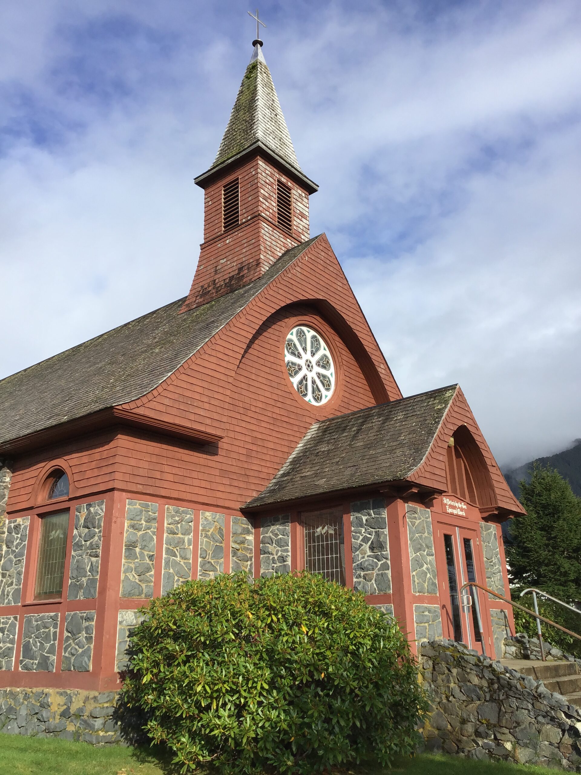 A historic church made of stone and cedar shake with a round, stained glass window above the entrance, and a tall steeple set against blue skies with light clouds