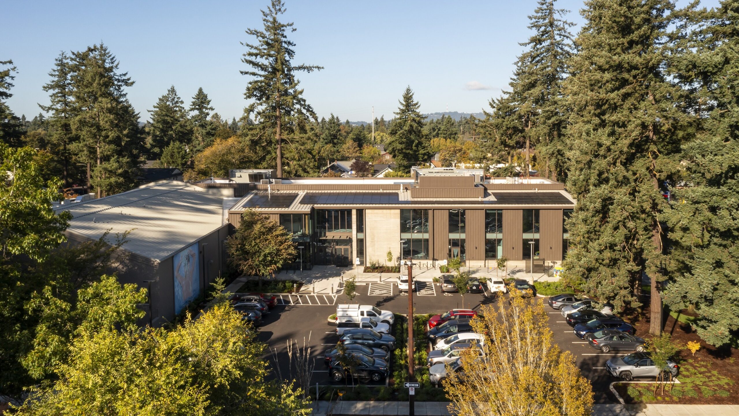 An aerial view shows Mt. Scott Community Center nestled among mature trees, with rooftop solar panels and a landscaped parking area.