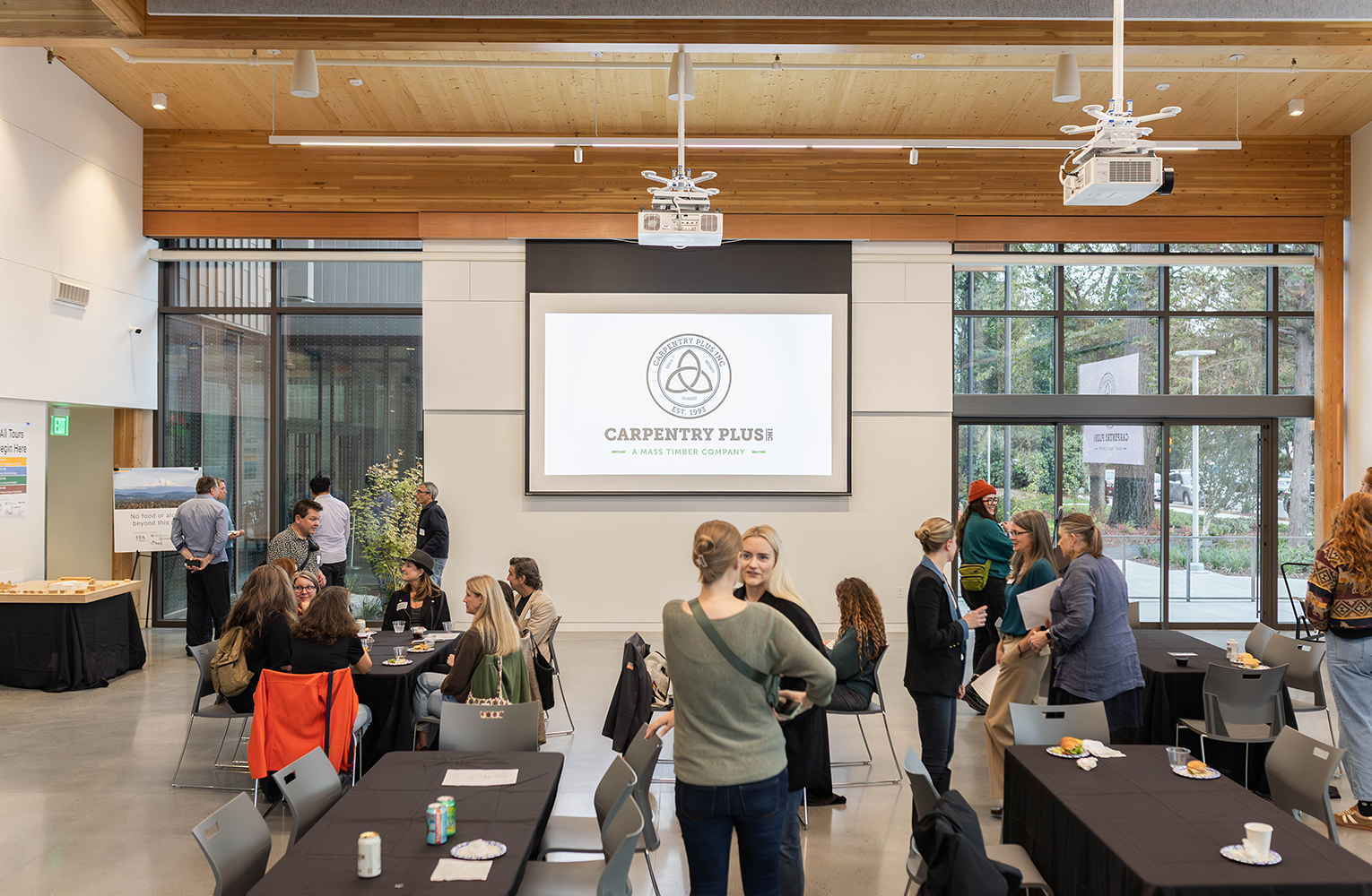 Community members gather for a public event in the event hall, meeting beneath exposed mass timber beams and integrated audiovisual systems