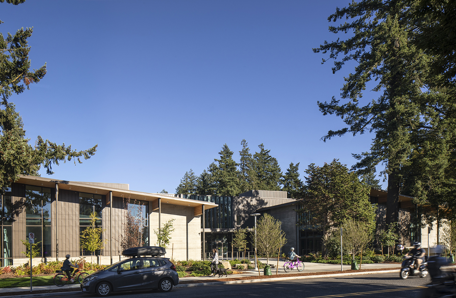 Community members walk and bike past the mass-timber Mt. Scott Community Center, framed by tall evergreens along SE 72nd Avenue.