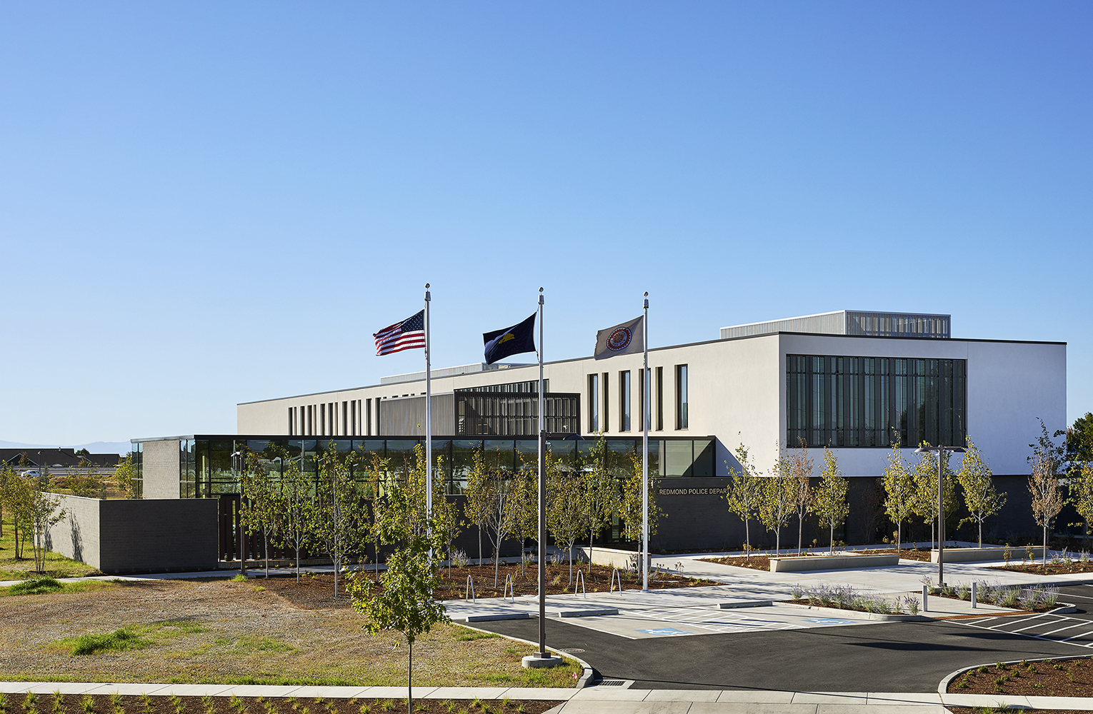 Exterior of the Redmond Public Safety Facility with three flagpoles in front, landscaped plaza, and a modern civic building featuring glass and metal façade elements.