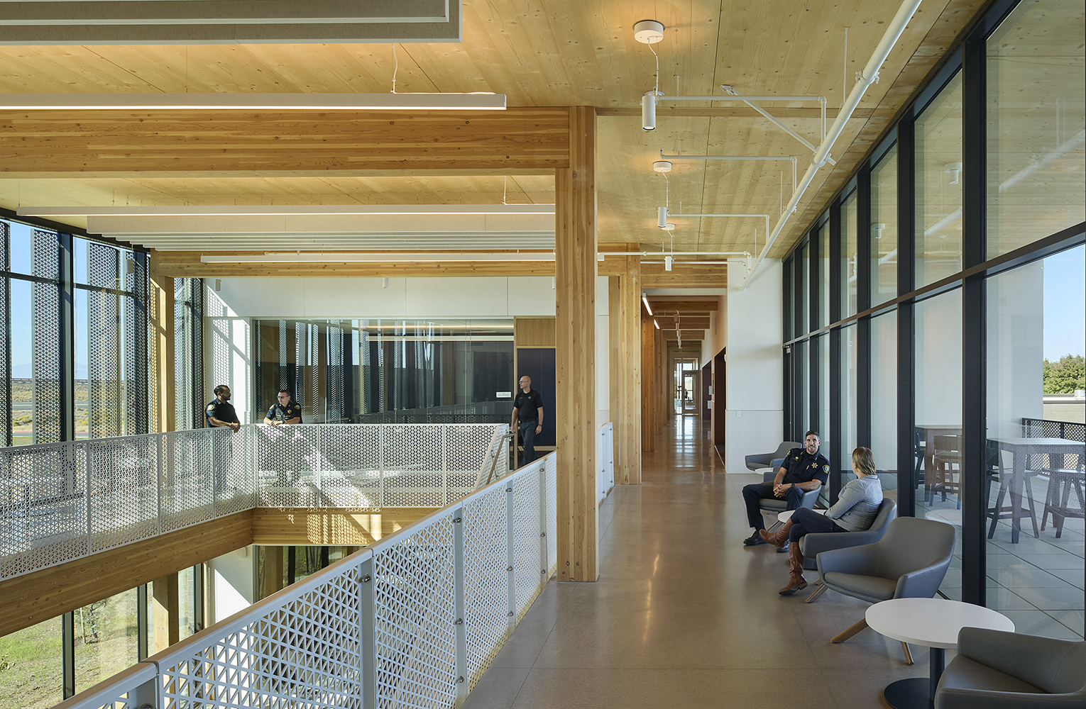 Daylit interior corridor at the Redmond Public Safety Facility with exposed mass timber structure, perforated metal screens filtering sunlight, and officers meeting and relaxing in lounge seating