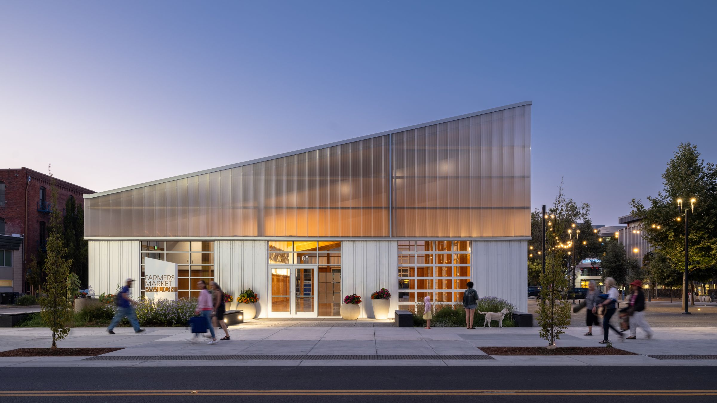 Visitors walk past the illuminated Eugene Farmers Market Pavilion at dusk, where a translucent façade reveals the warm timber structure within.